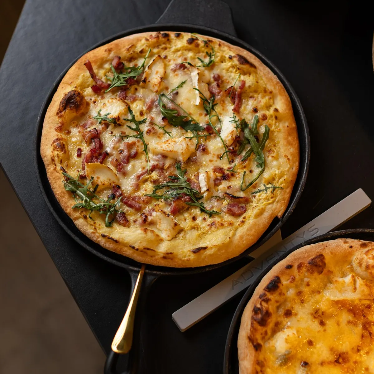 Deux pizzas dans des poêles à pizza sur une table avec une bouteille de sauce piquante en arrière-plan
