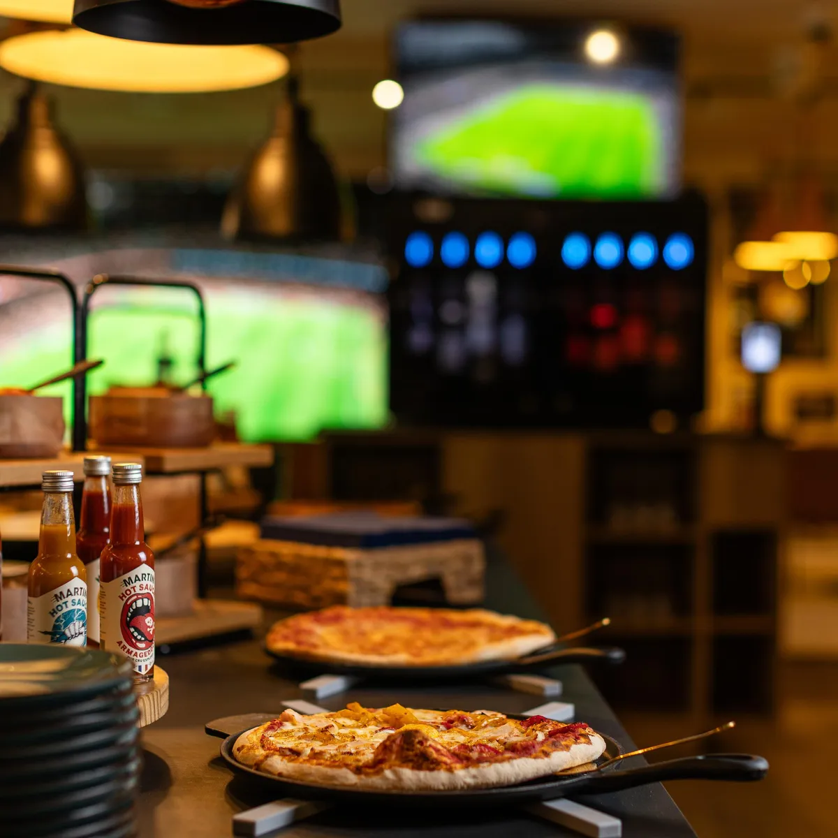 Deux pizzas sur des plaques en métal posées sur une table dans un restaurant avec des écrans diffusant un match de football en arrière-plan