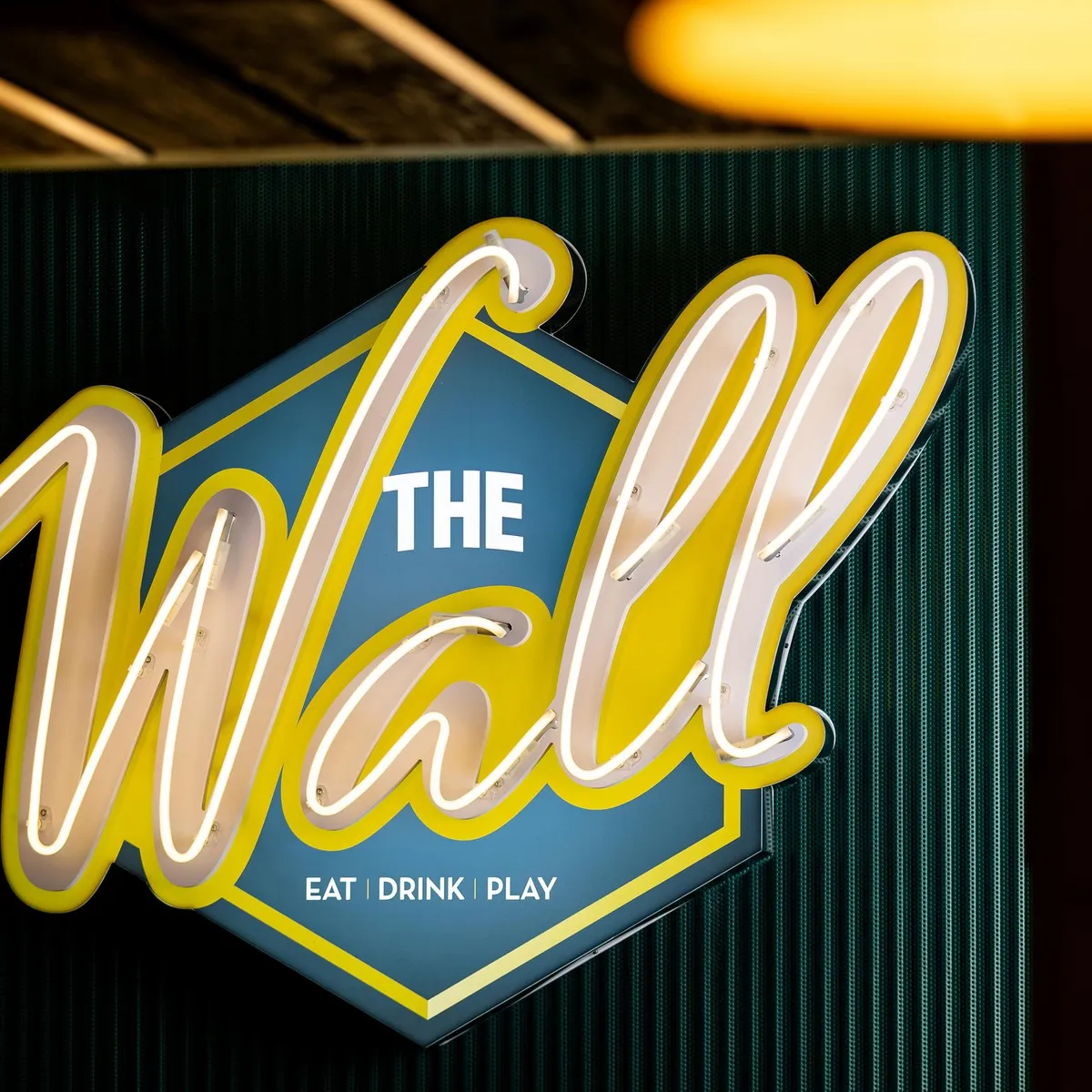 Enseigne lumineuse avec l'inscription "The Wall" et les mots "Eat Drink Play" en dessous, fixée à un mur sombre