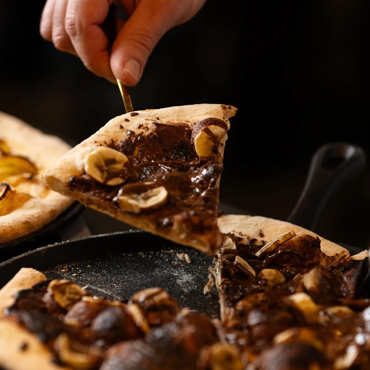 Une main tient une tranche de pizza aux champignons et aux noix, tandis que deux pizzas sont posées sur des plaques de cuisson