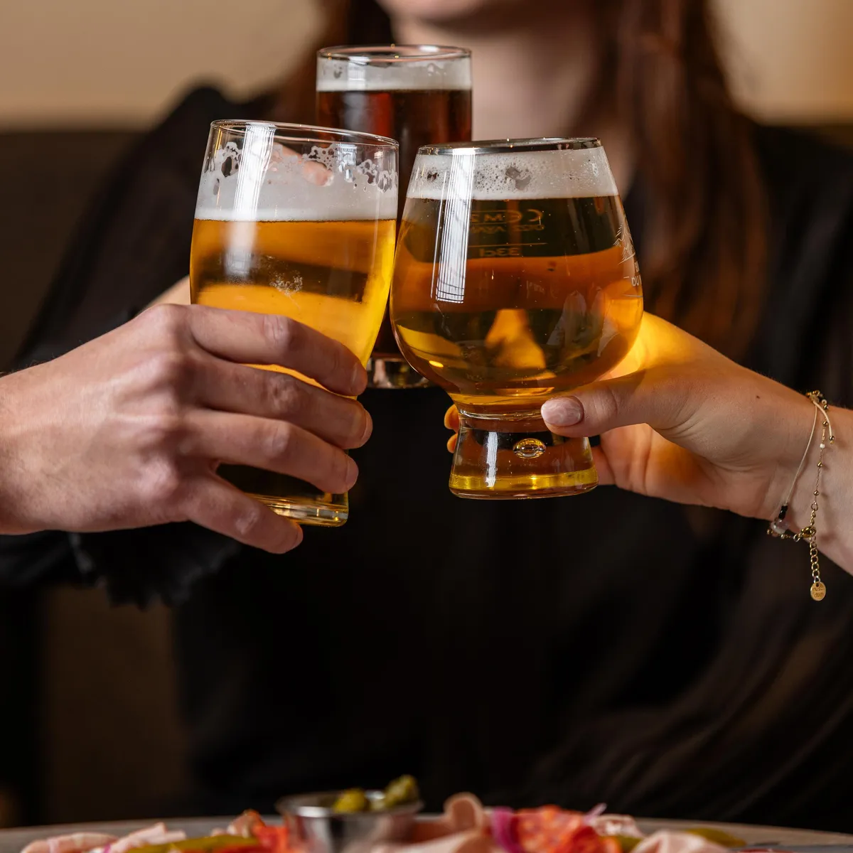 Deux personnes lèvent des verres de bière en se faisant face, avec une assiette de charcuterie et de fromage sur la table devant eux
