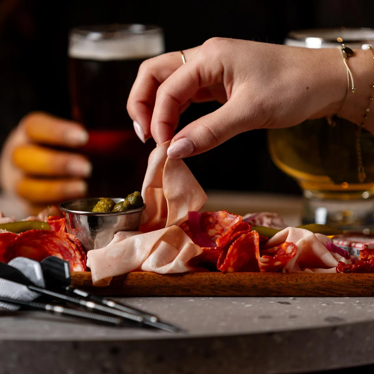 Une personne prend un morceau de charcuterie sur une planche avec des cornichons et des olives, accompagnée de verres de bière