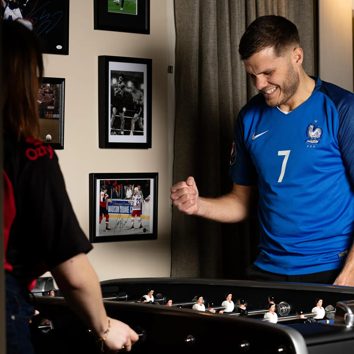 Un homme en maillot de football bleu joue au baby-foot avec deux femmes dans une pièce décorée de photos encadrées