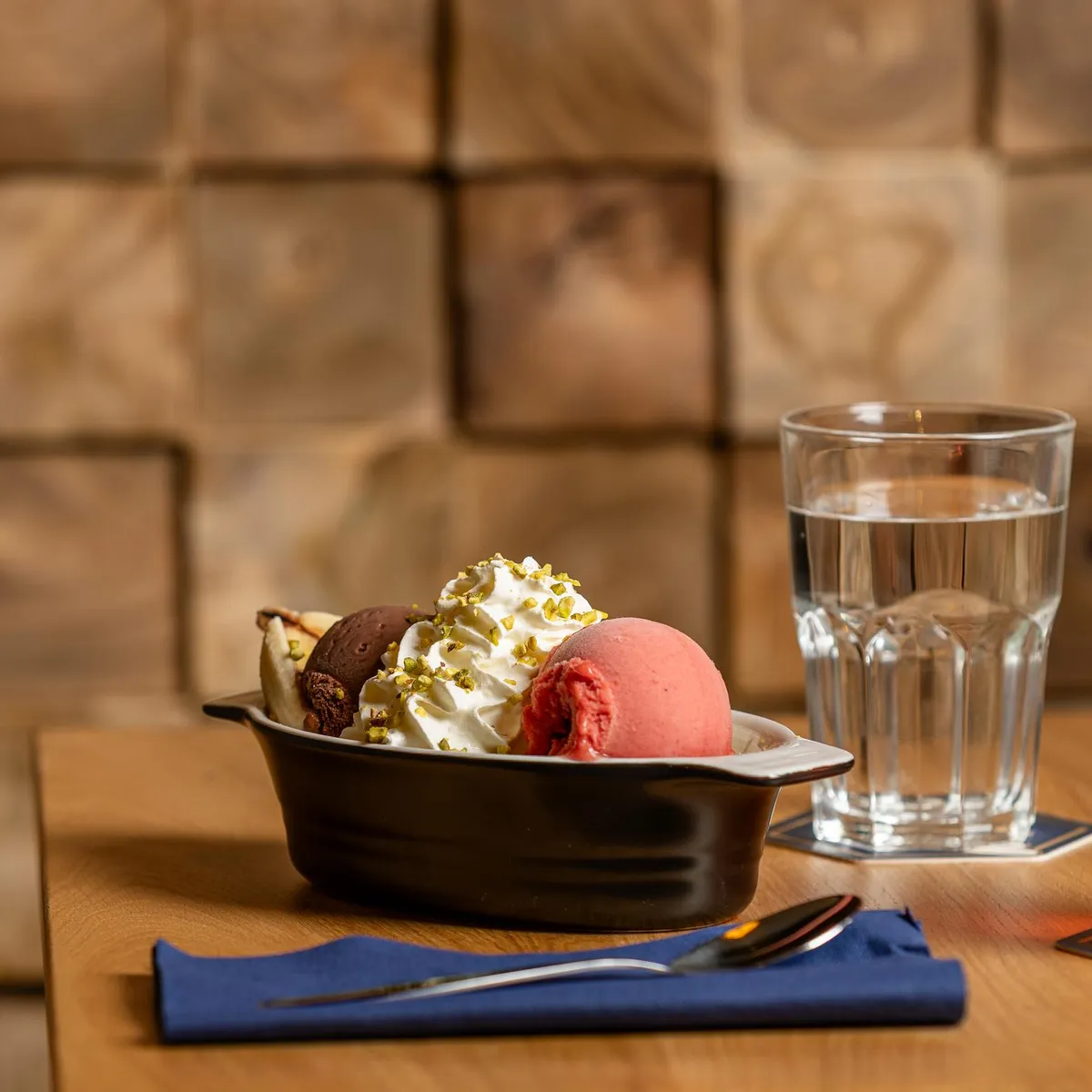 Un bol d'accompagnement avec des glaces et une garniture, un verre d'eau et un verre de boisson rouge sur une table en bois devant un mur de briques