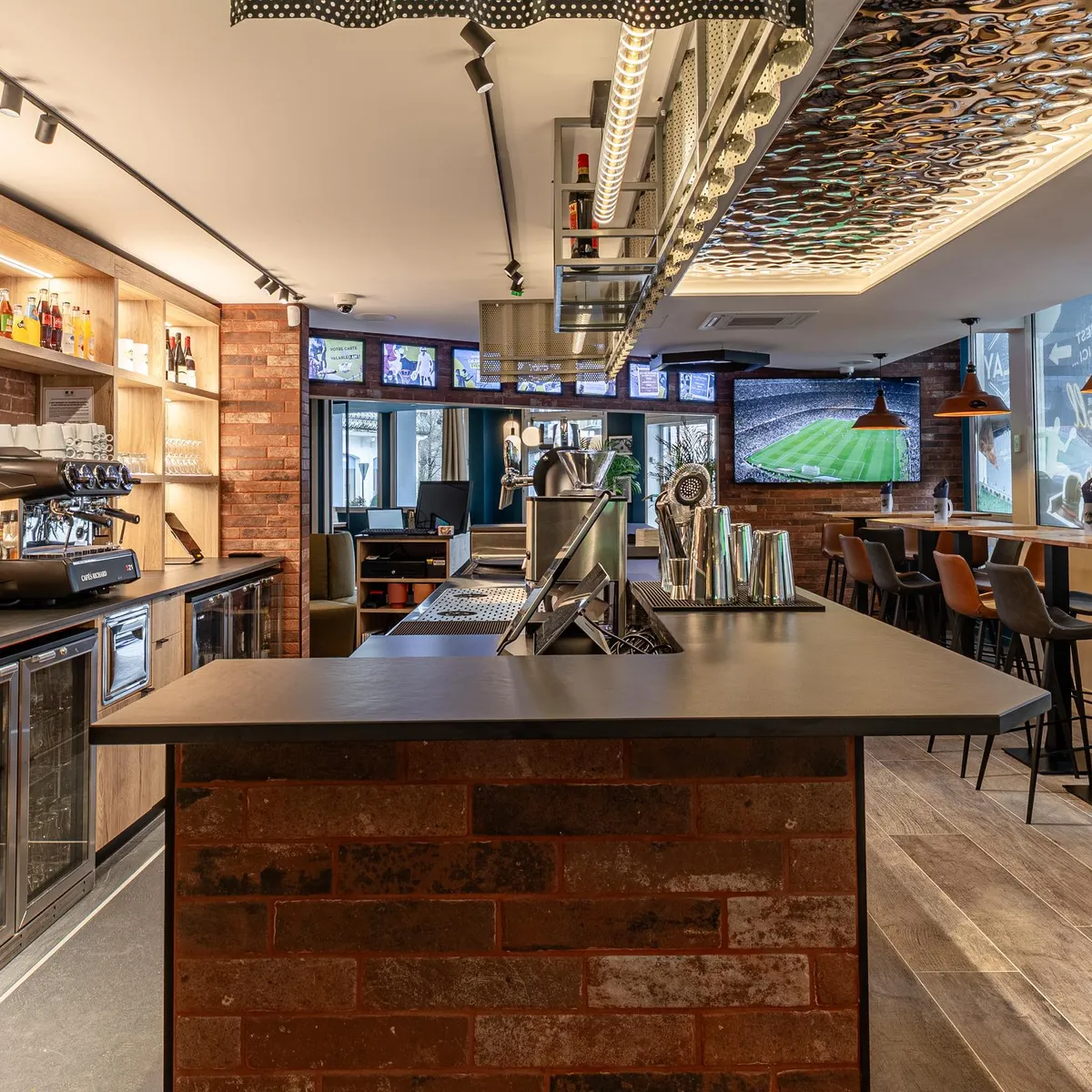 Bar avec comptoir central, écrans diffusant un match de football, étagères garnies de bouteilles et tabourets de bar