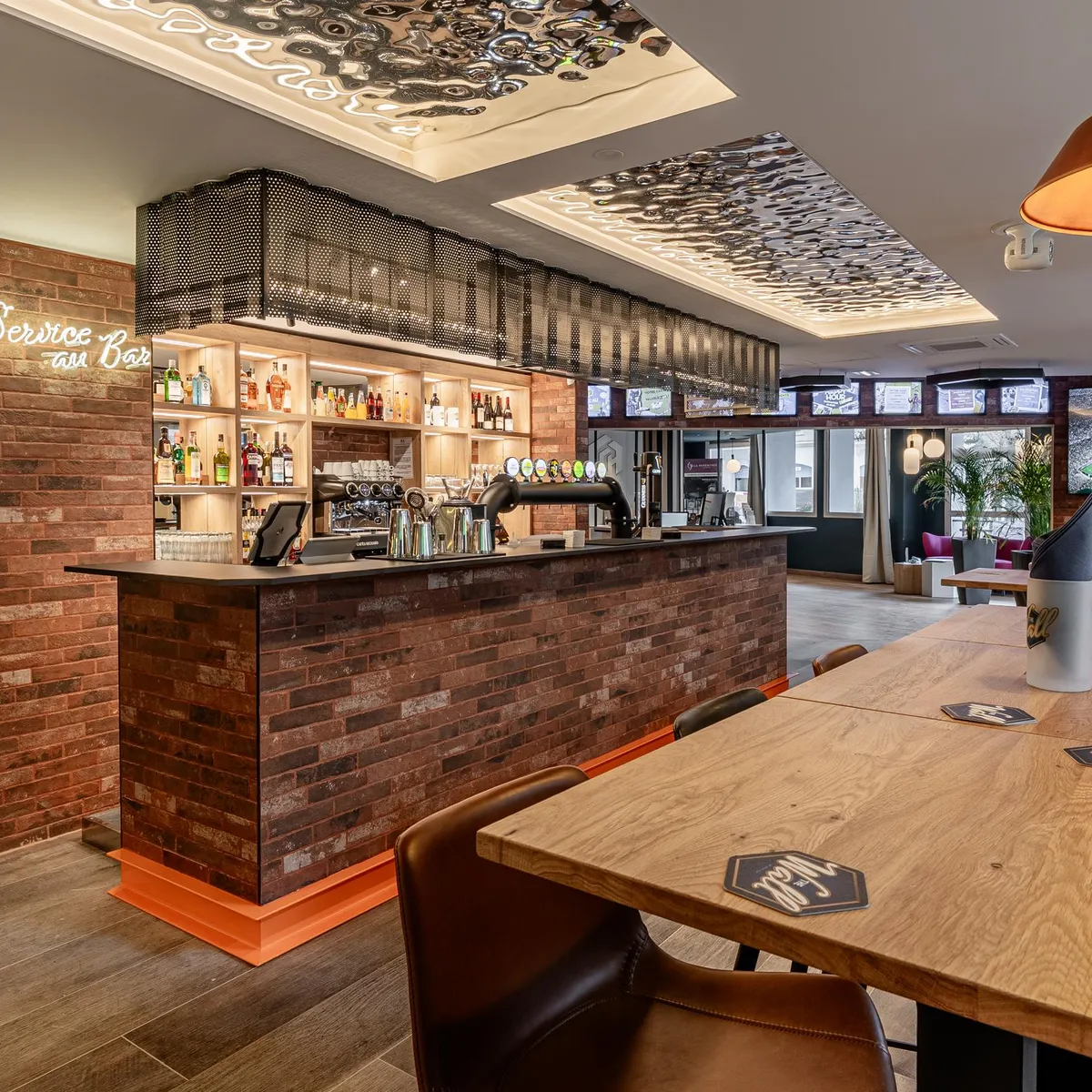 Bar avec comptoir en briques, étagères à bouteilles et tables en bois, avec des écrans diffusant un match de football en arrière-plan