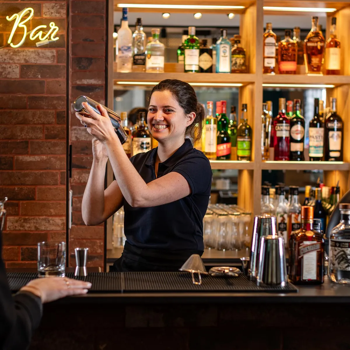 Une barmaid prépare un cocktail derrière un bar bien fourni en bouteilles et spiritueux
