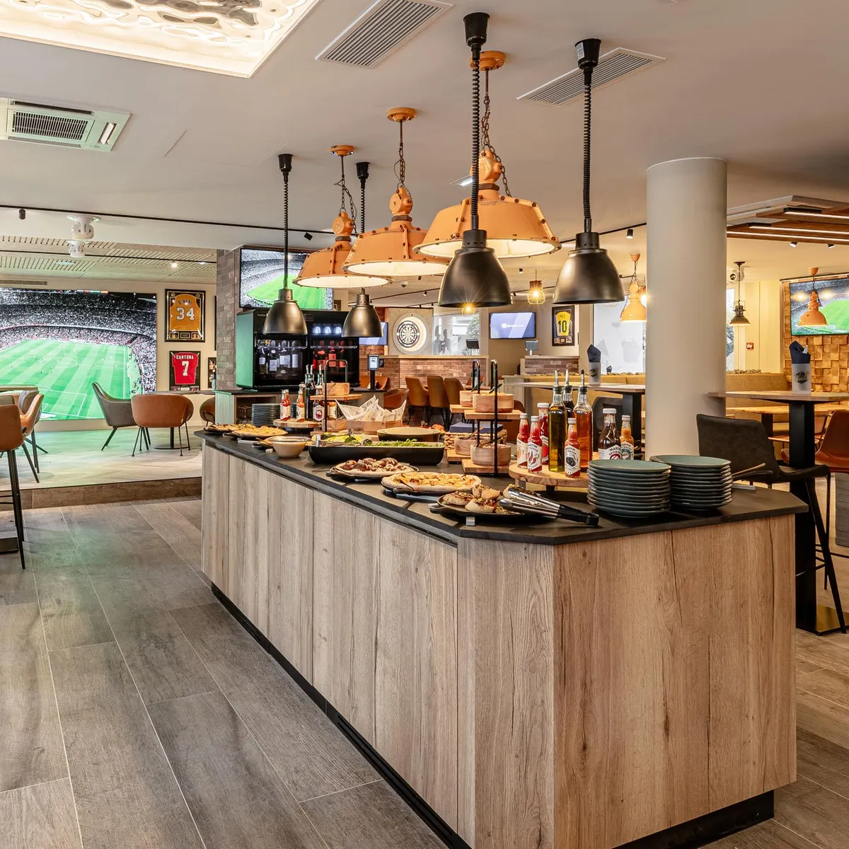 Un bar avec un comptoir en bois clair, des tabourets et des écrans diffusant un match de football, avec des boissons et de la nourriture disposés sur le comptoir