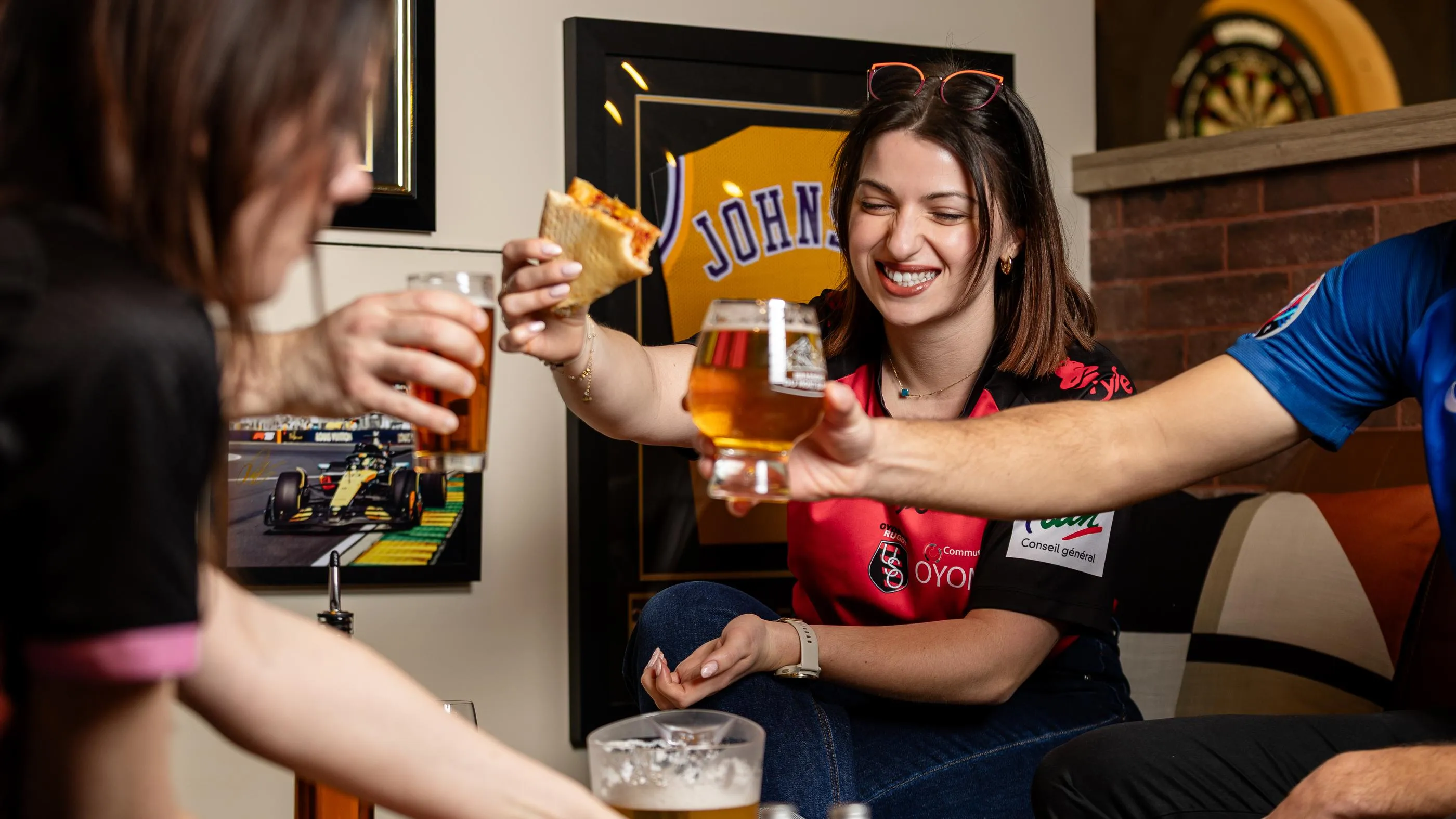 Une femme sourit en tenant une part de pizza et un verre de bière, entourée de personnes assises autour d'une table