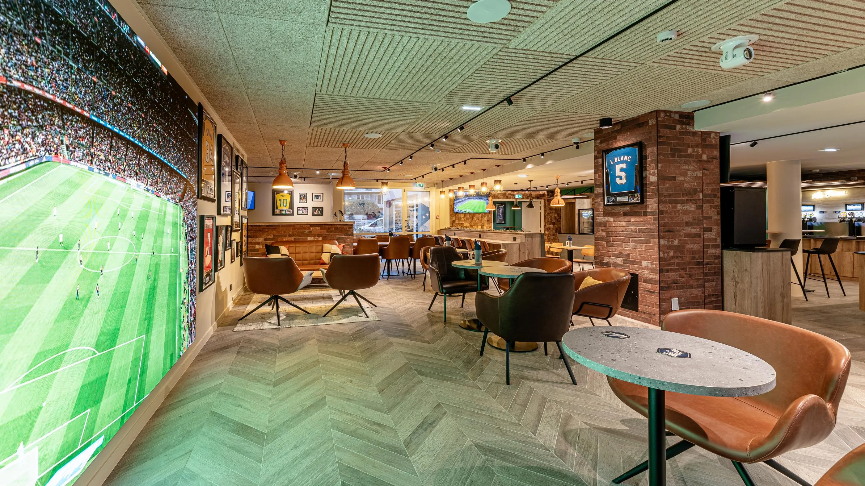 Un espace intérieur avec des sièges, des tables et un grand écran affichant un terrain de football, entouré de décorations sportives