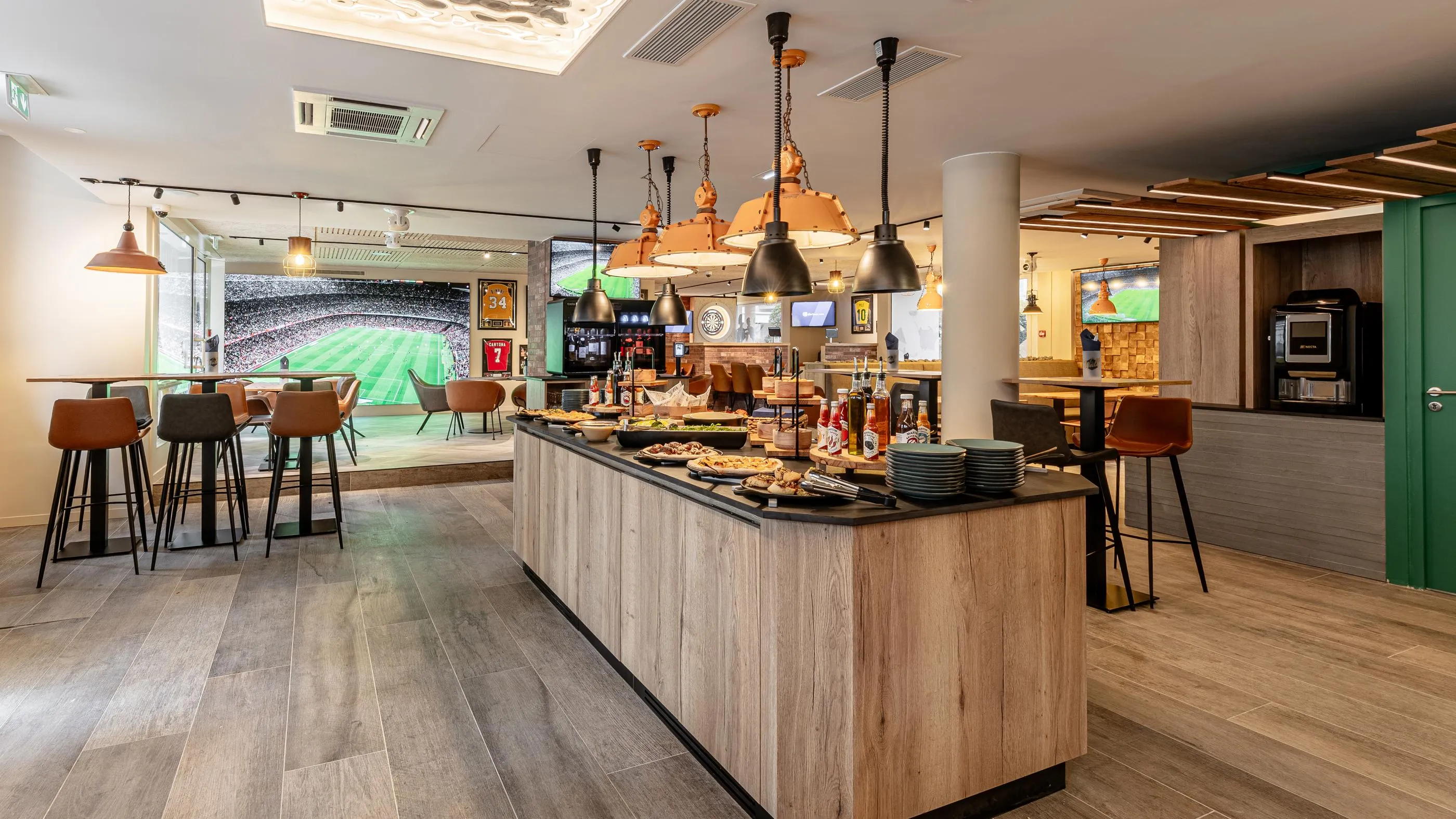 Un bar avec un comptoir en bois clair, des tabourets et des écrans diffusant un match de football, avec des boissons et de la nourriture disposés sur le comptoir