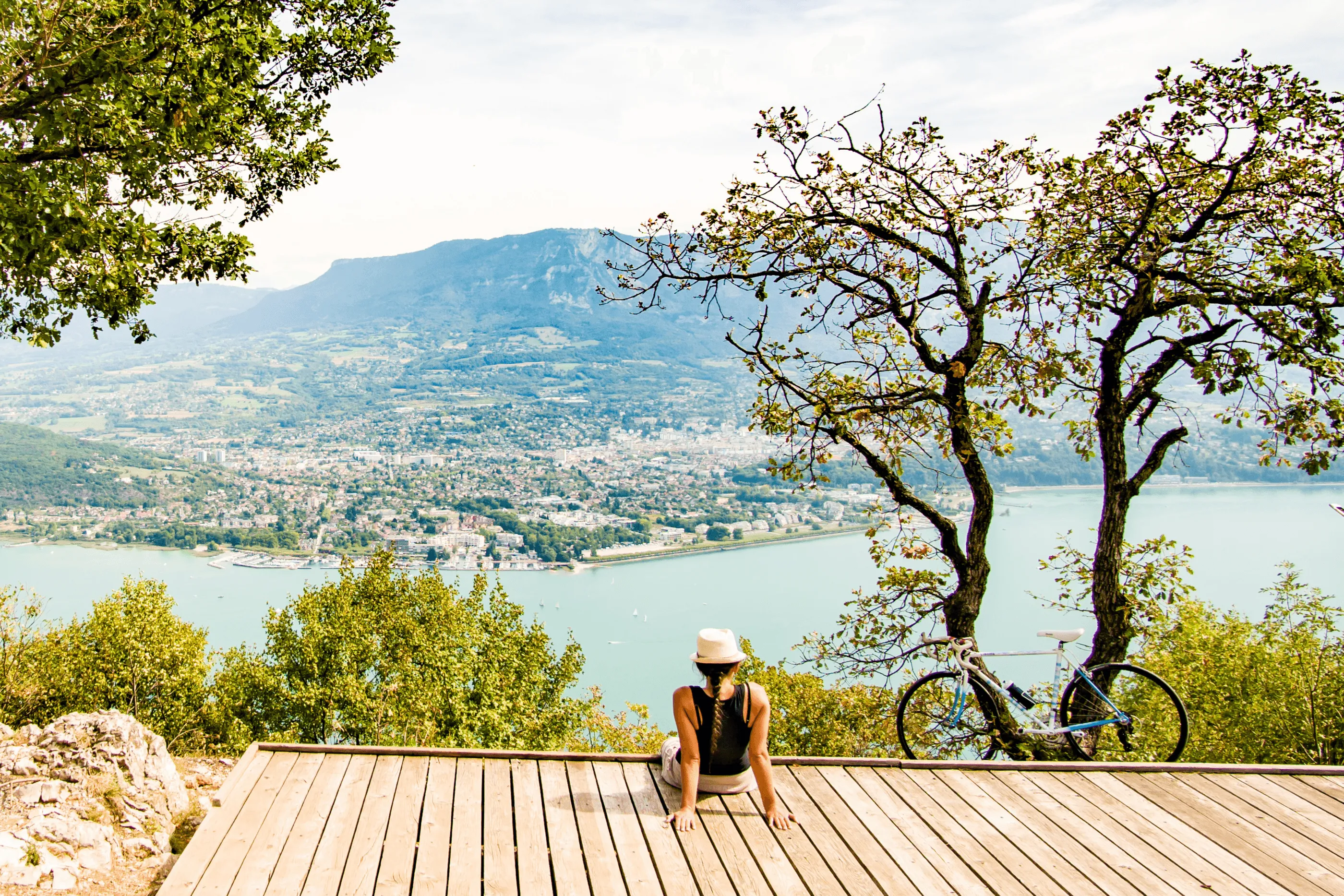 Une personne portant un chapeau et un haut noir est assise sur une plateforme en bois avec un vélo à côté d'elle, face à un lac et une ville entourée de montagnes