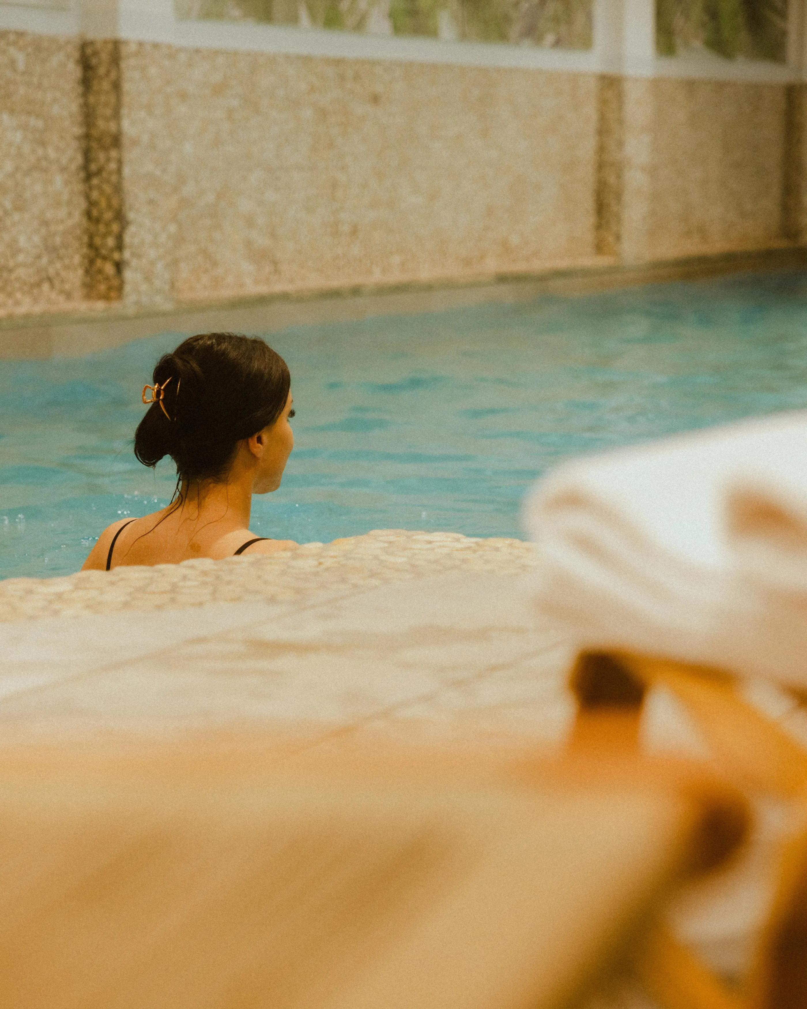 Une personne avec les cheveux attachés est assise dans une piscine, vue de dos, avec un mur en pierre en arrière-plan