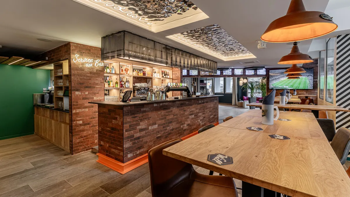 Bar avec comptoir en briques, étagères à bouteilles et tables en bois, avec des écrans diffusant un match de football en arrière-plan