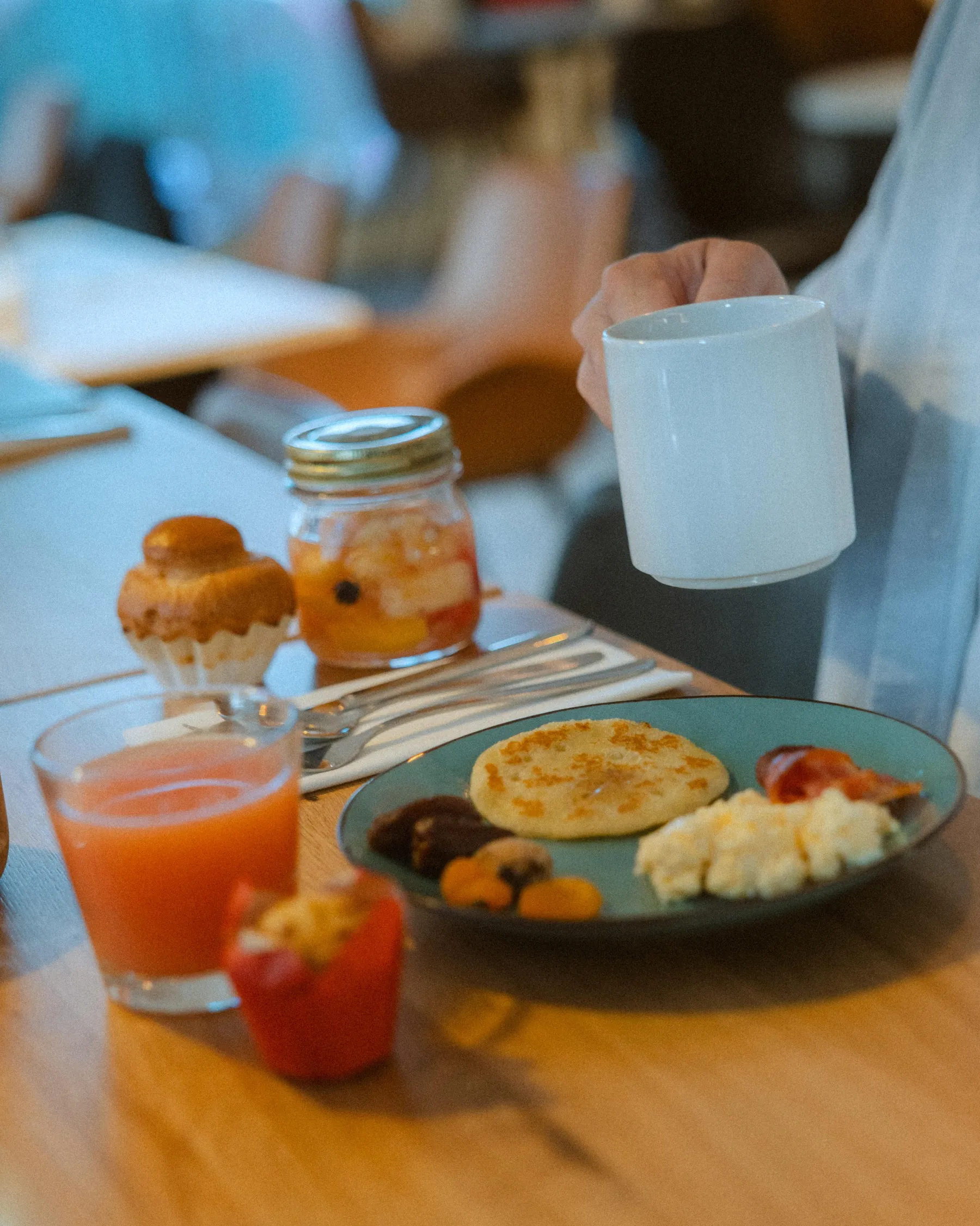 Une personne tient une tasse blanche à café, avec une assiette de nourriture et un verre de jus d'orange sur une table