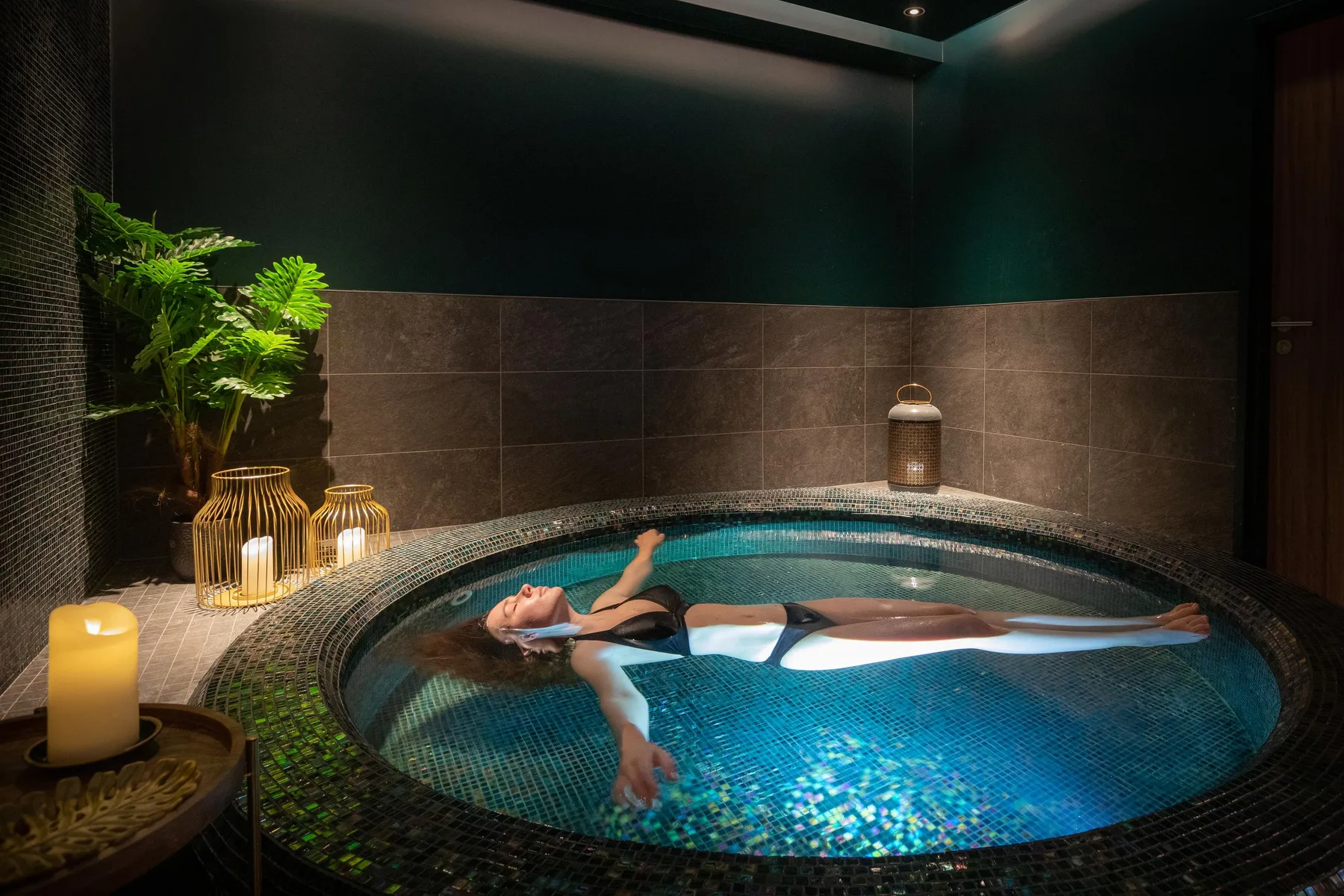 Une personne allongée dans un bain à remous entouré de bougies et de plantes vertes dans une pièce sombre