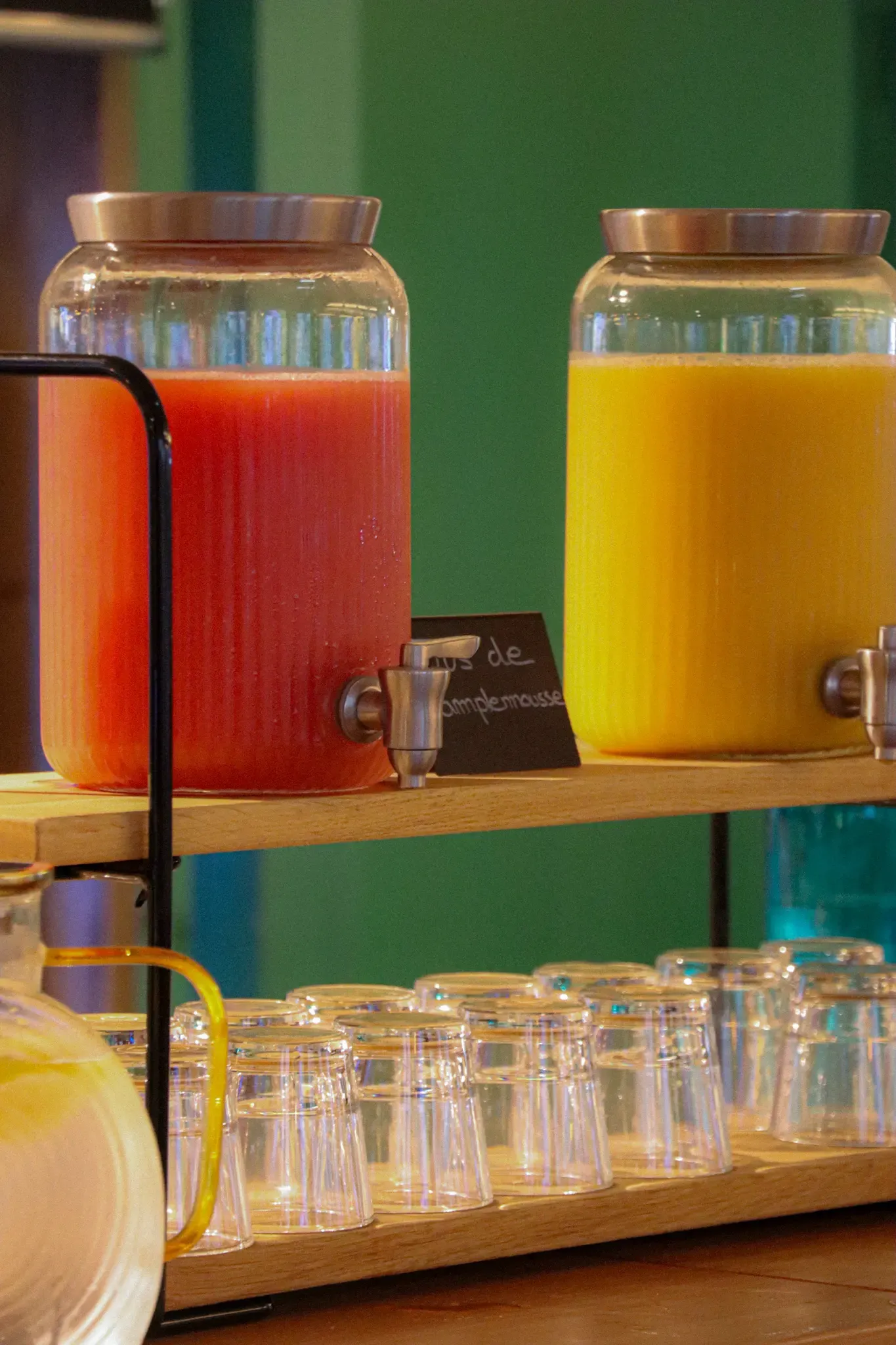 Deux grands récipients transparents contenant des boissons de couleurs rouge et orange sont posés sur une étagère en bois avec des verres en dessous