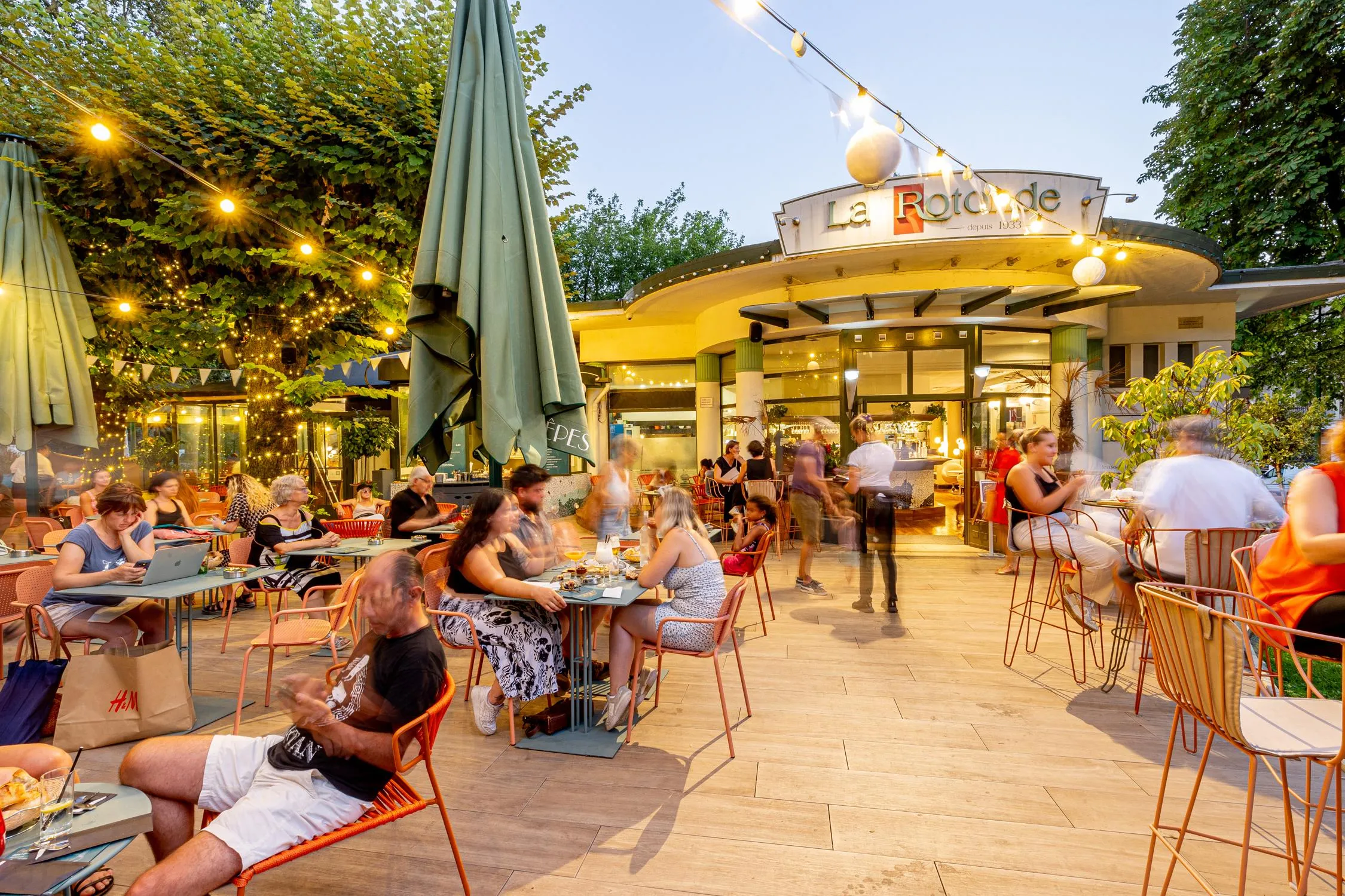 Terrasse de restaurant animée avec des clients assis à des tables sous des parasols, entourée d'arbres et de lumières décoratives, devant un bâtiment portant l'enseigne "La Rotonde"