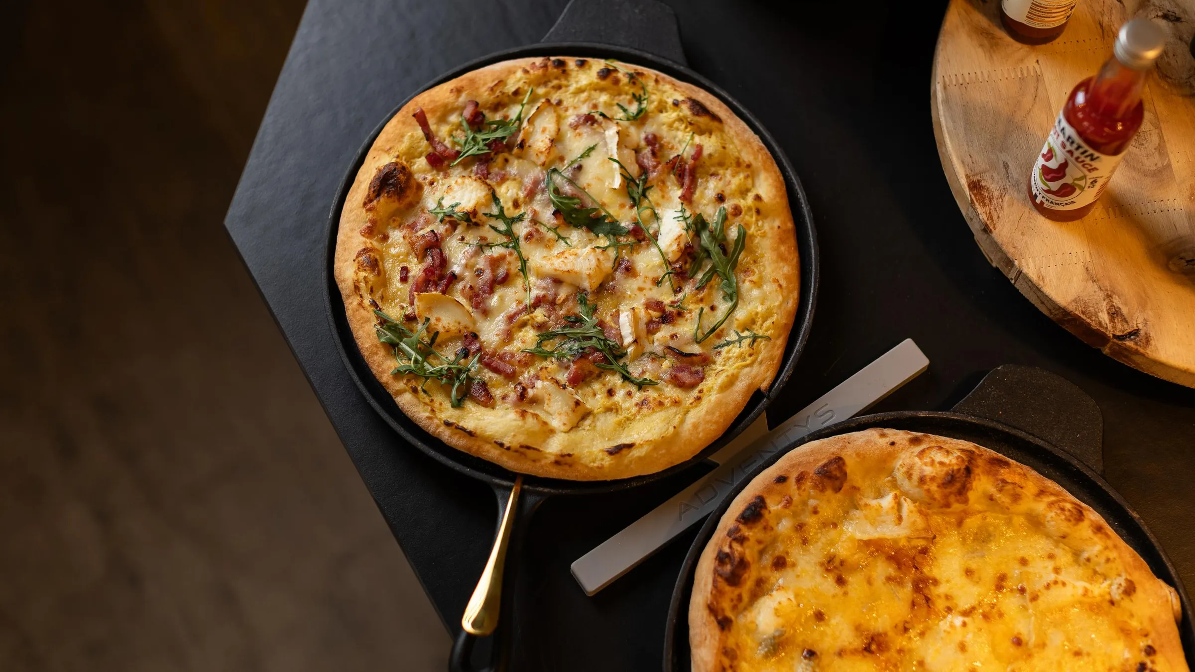 Deux pizzas dans des poêles à pizza sur une table avec une bouteille de sauce piquante en arrière-plan