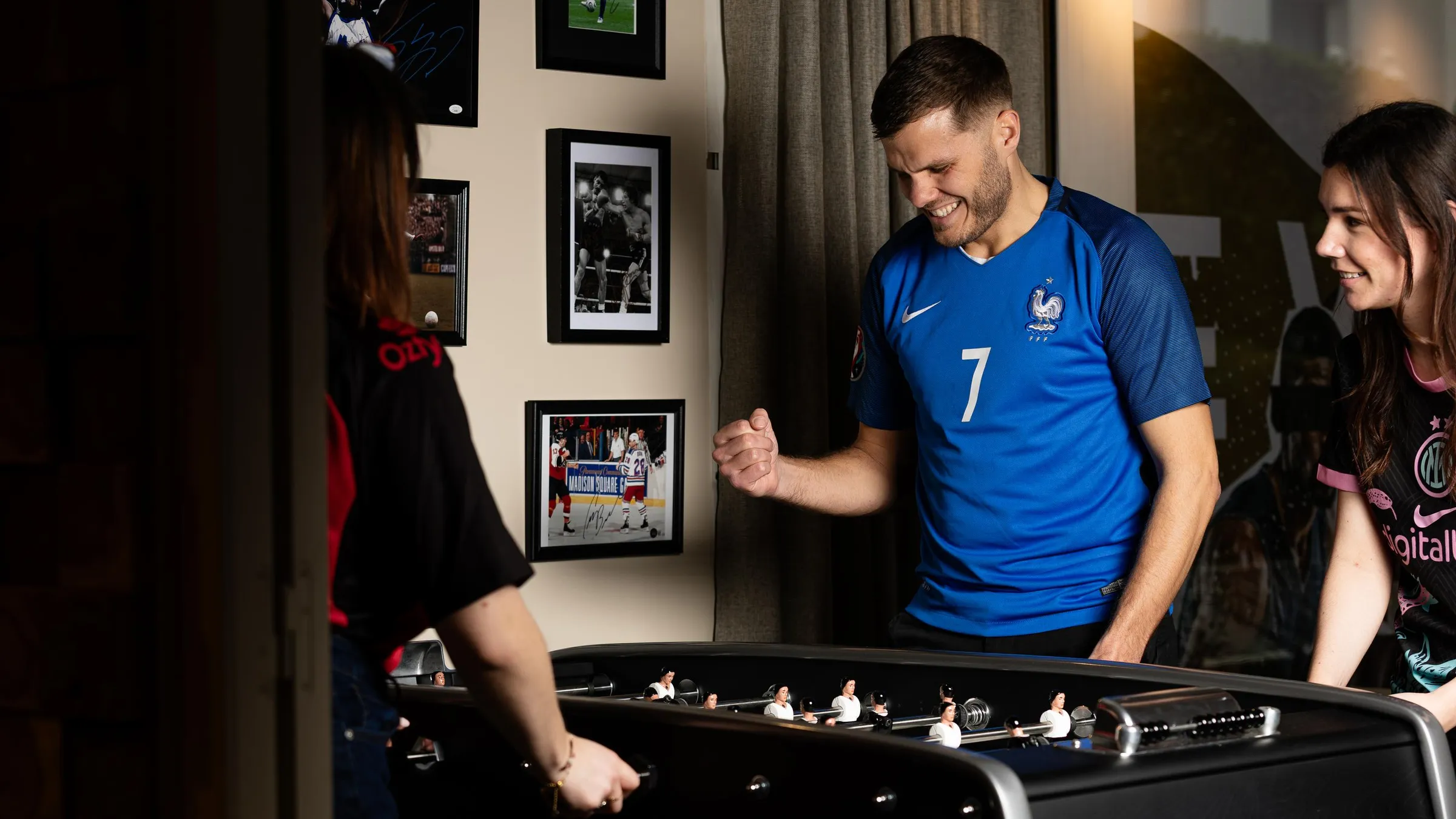Un homme en maillot de football bleu joue au baby-foot avec deux femmes dans une pièce décorée de photos encadrées