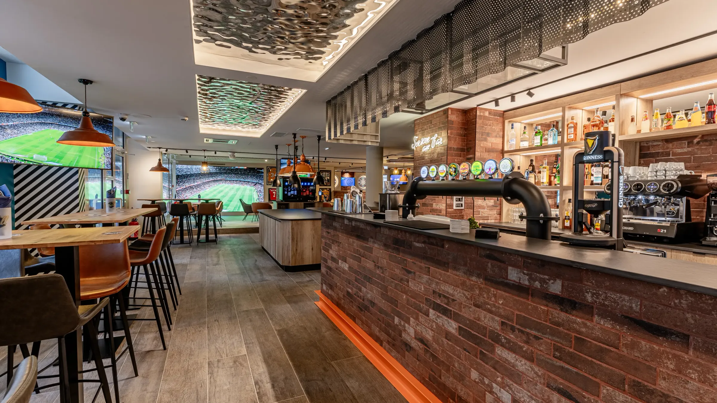 Bar avec comptoir en brique, tables et chaises, écrans diffusant un match de football, étagères remplies de bouteilles et de verres