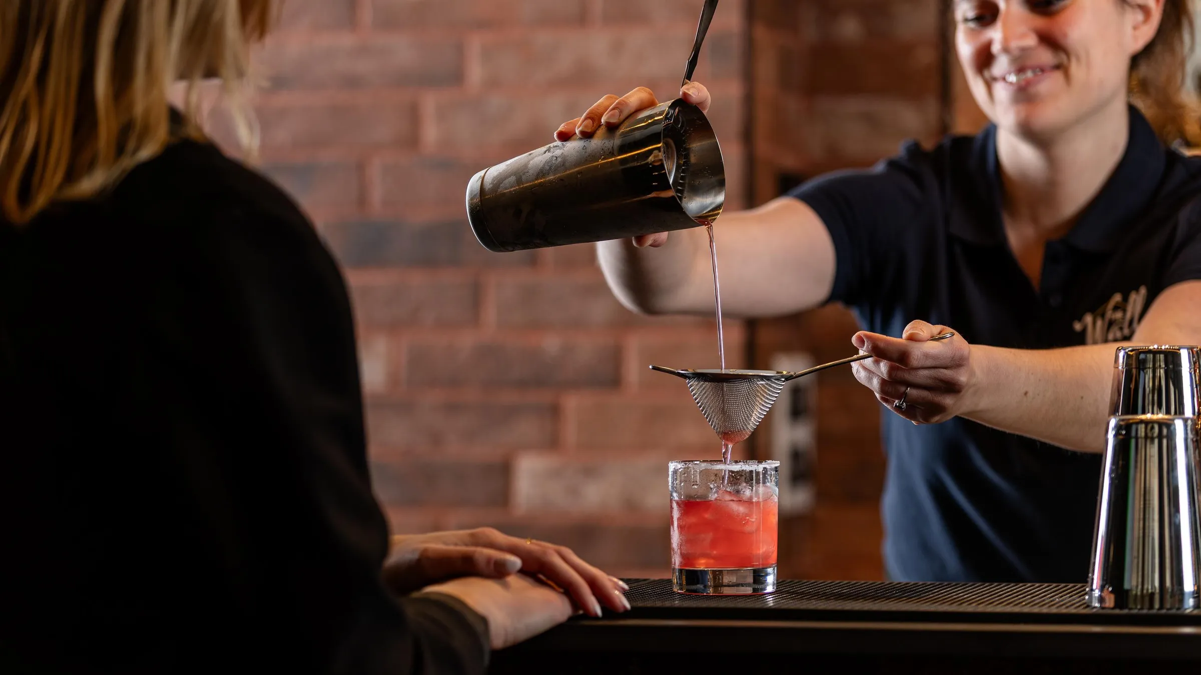 Une personne verse un liquide à travers une passoire dans un verre contenant un cocktail rose