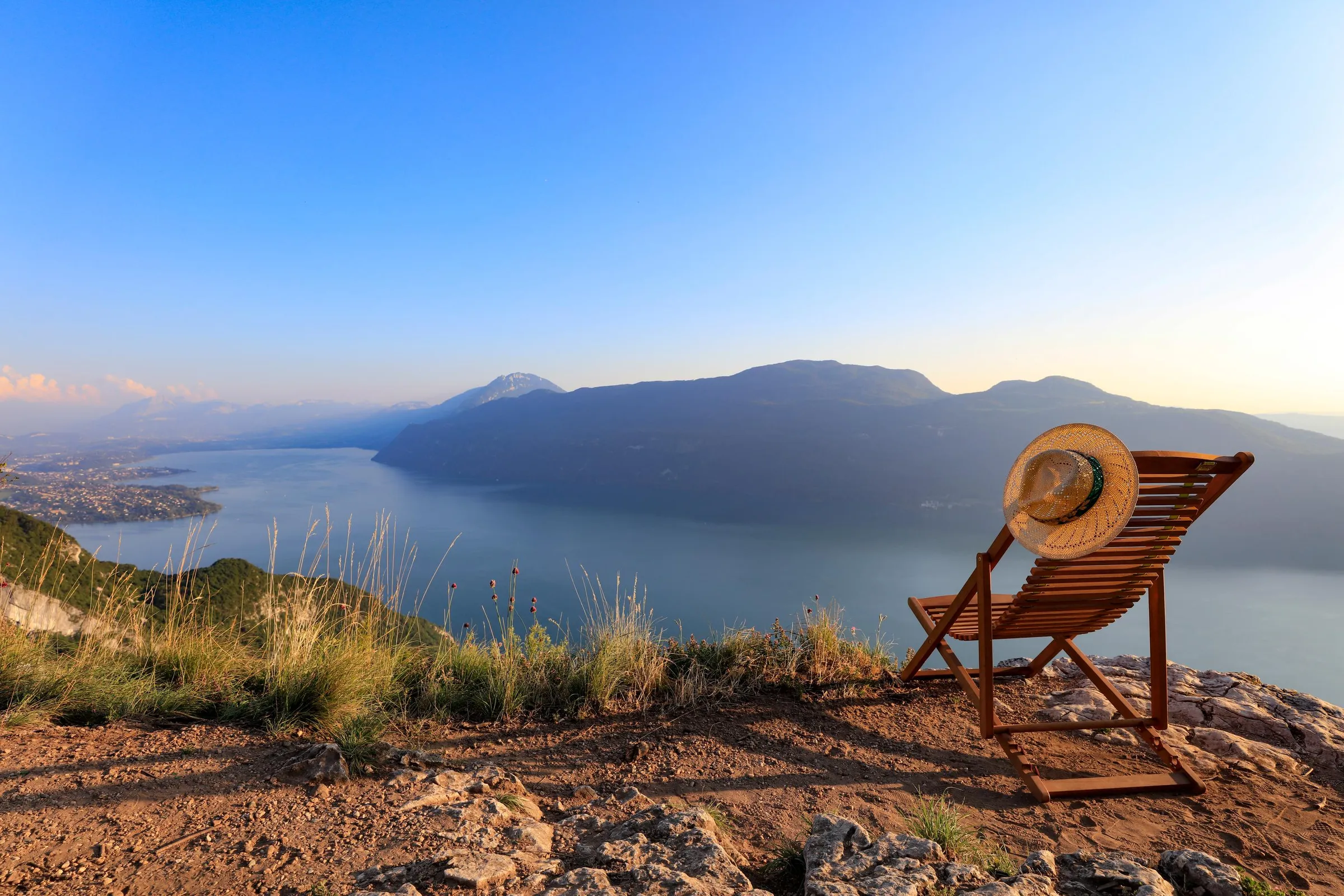 Une chaise longue avec un chapeau posée sur un terrain en pente face à un lac et des montagnes au loin