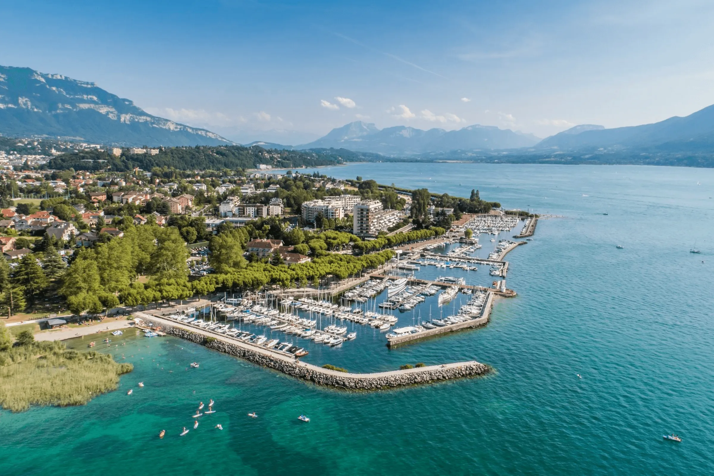 Village au bord d'un lac avec marina, montagnes en arrière-plan, et personnes en paddle dans l'eau