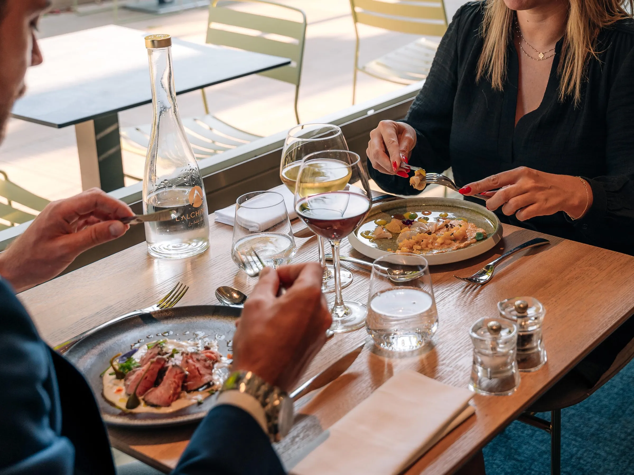 Trois personnes assises autour d'une table partagent un repas composé de plats variés, accompagnés de verres de vin et d'une bouteille d'eau