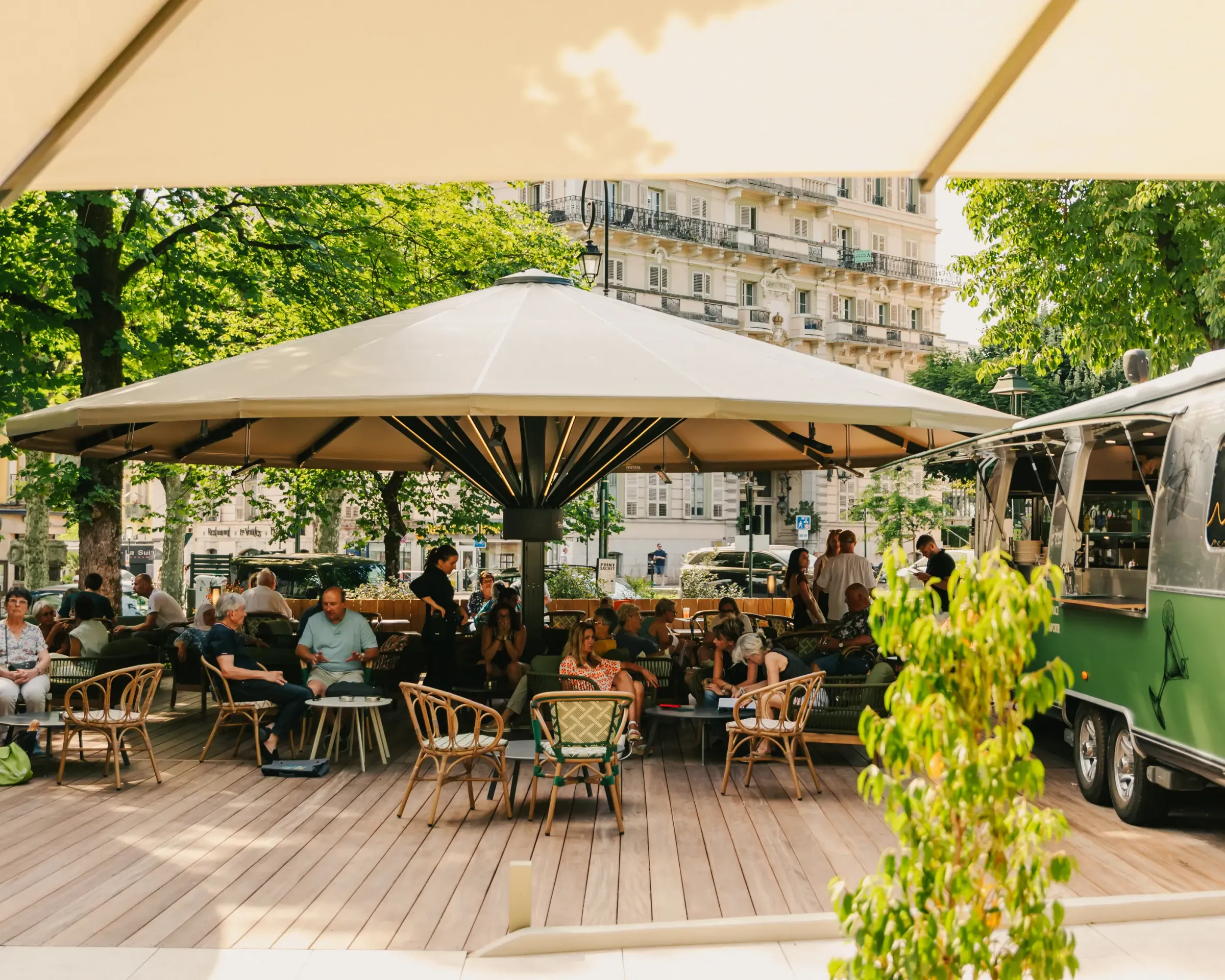 Un café en terrasse couvert avec des parasols, des clients assis à des tables sous les arbres, entouré de bâtiments et de verdure