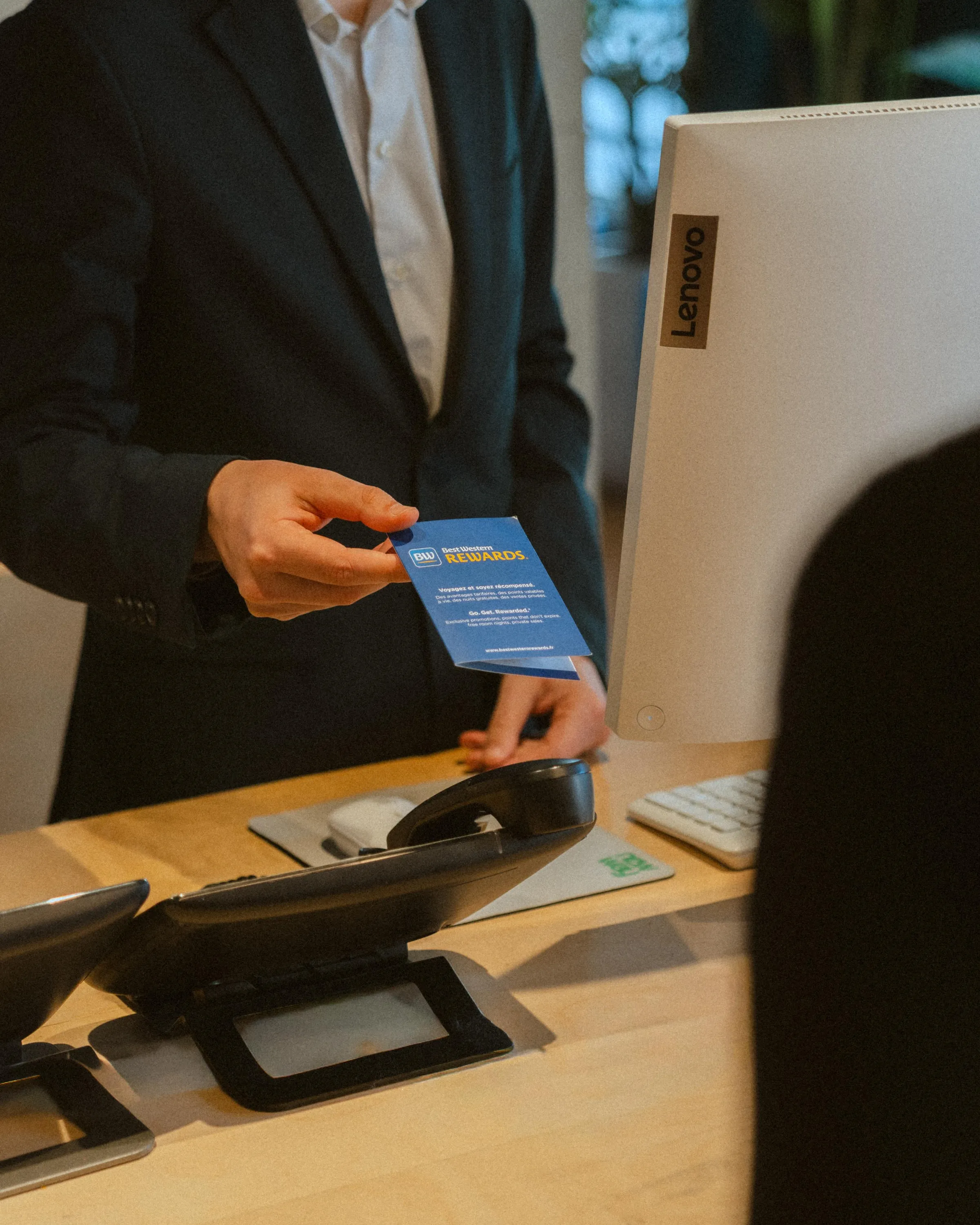 Une personne tend une carte de fidélité Best Western Rewards à un employé devant un ordinateur portable Lenovo
