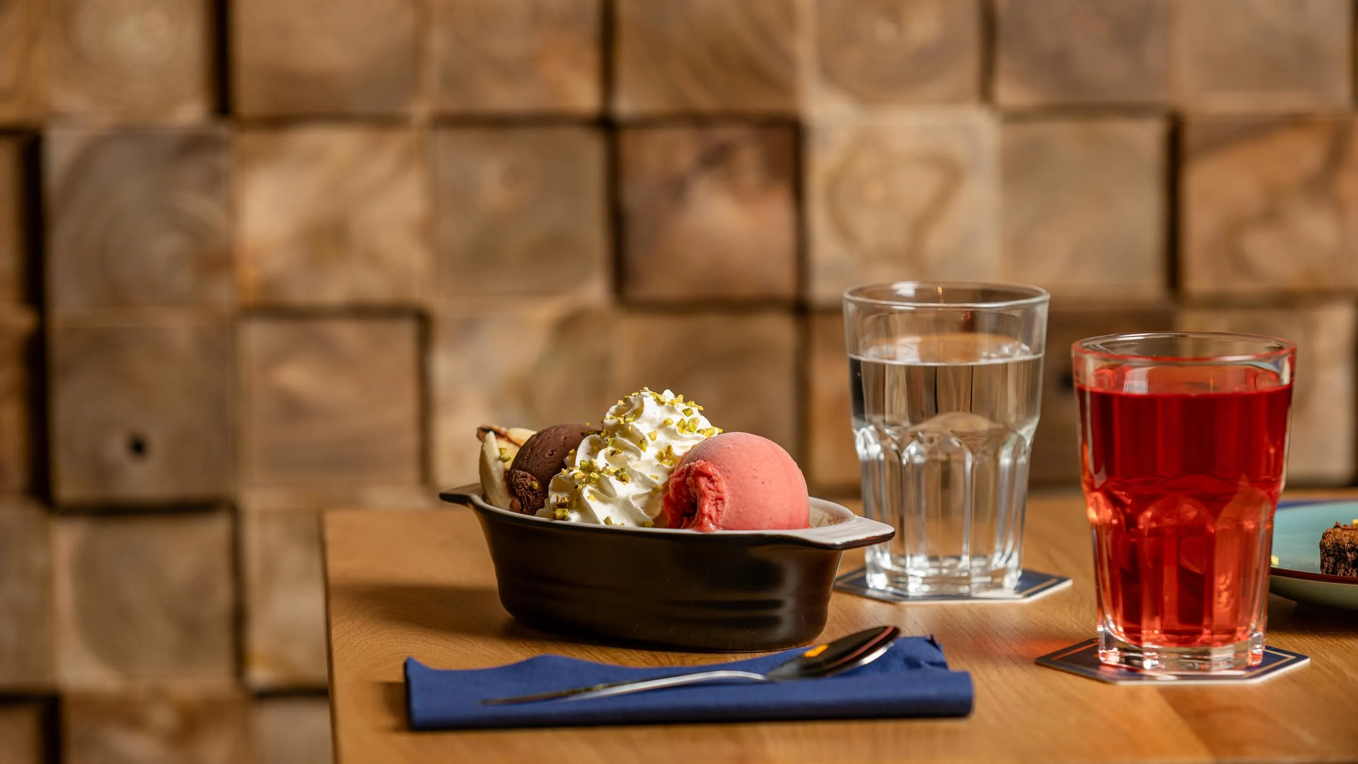 Un bol d'accompagnement avec des glaces et une garniture, un verre d'eau et un verre de boisson rouge sur une table en bois devant un mur de briques