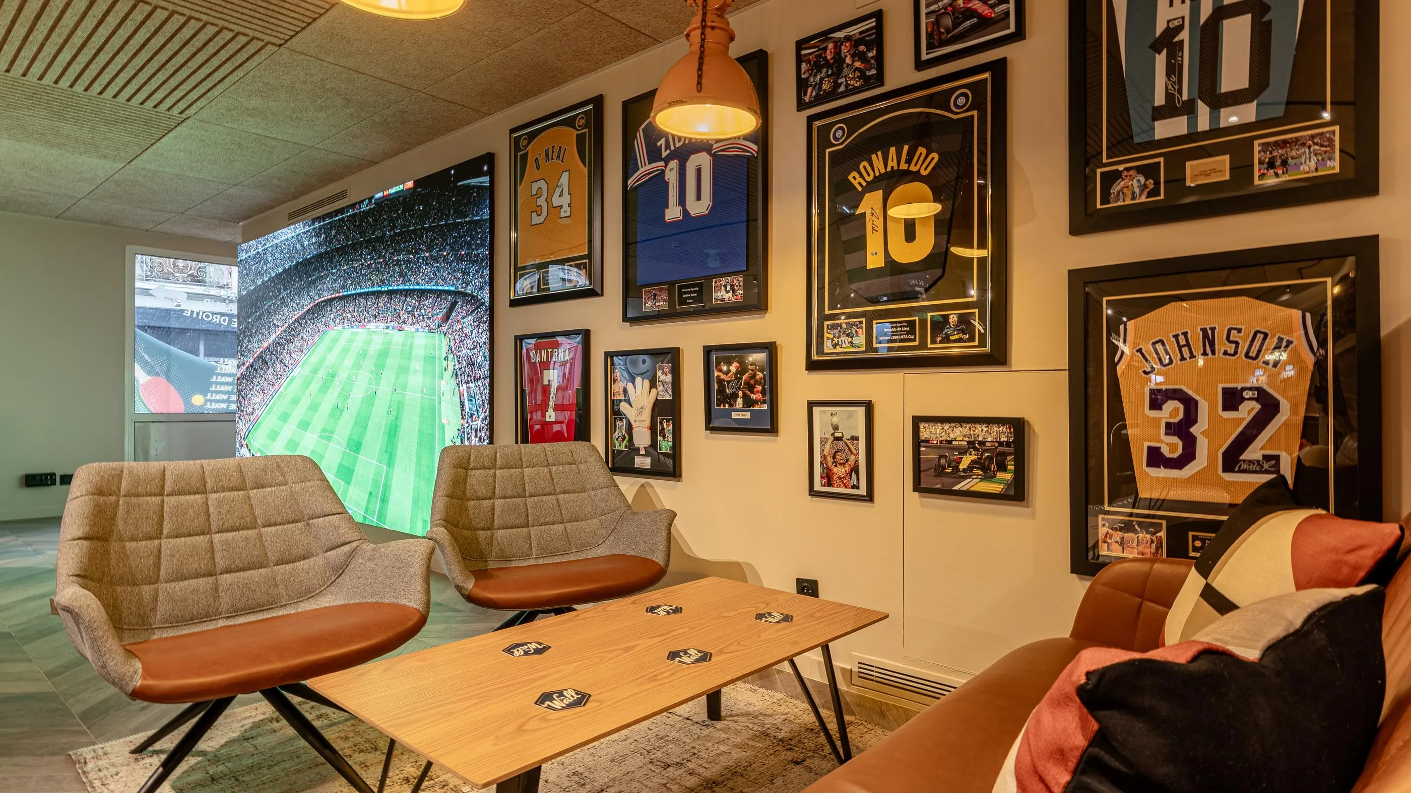 Un salon avec des chaises et une table basse, des maillots de sport encadrés aux murs et une télévision affichant un match de football