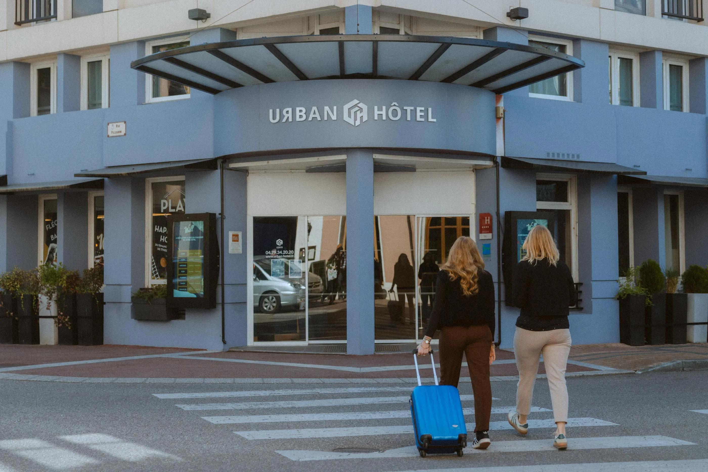 Deux personnes poussant un chariot de valise bleue traversent un passage piéton devant l'entrée d'un hôtel nommé Urban Hôtel