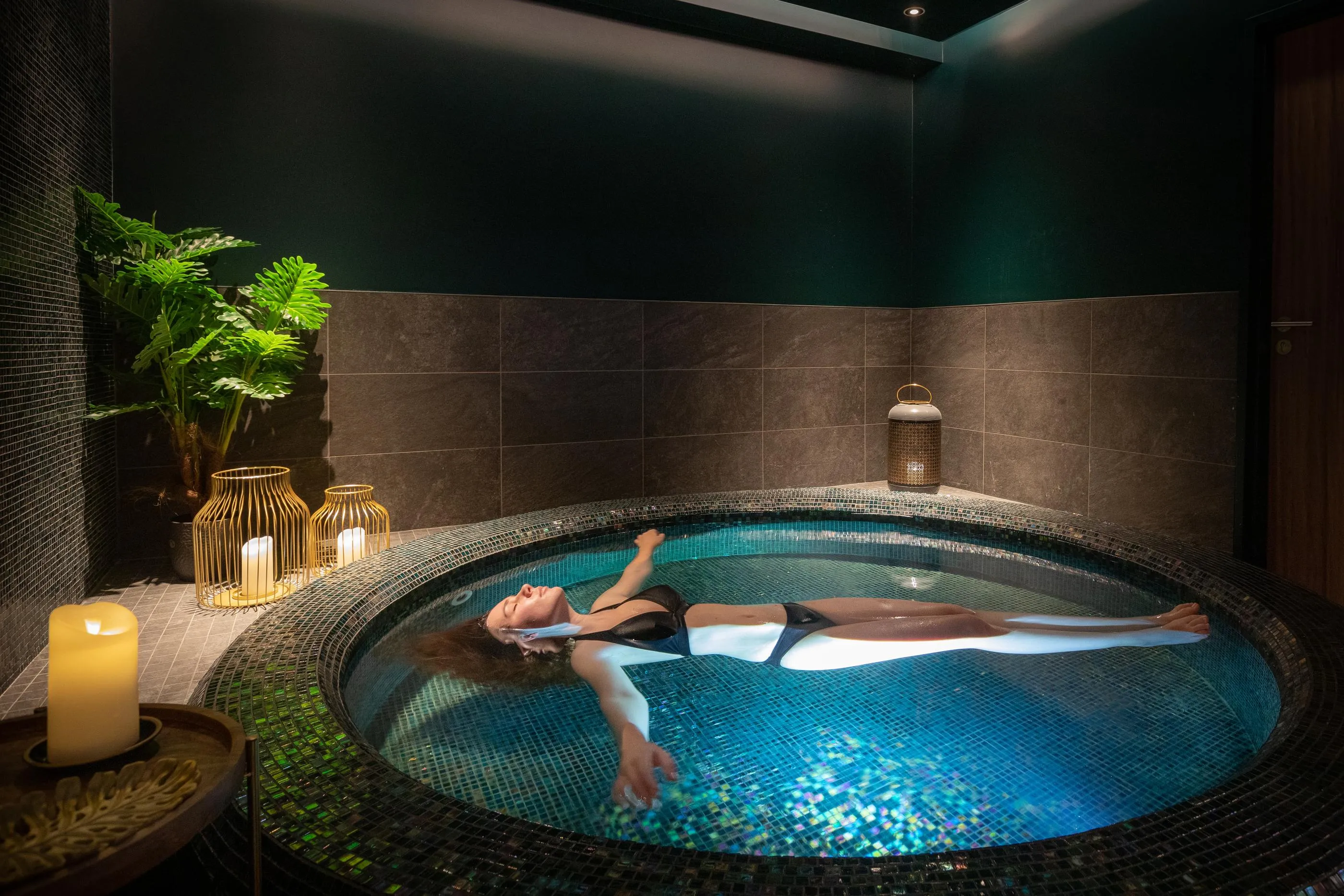 Une personne allongée dans un bain à remous entouré de bougies et de plantes vertes dans une pièce sombre