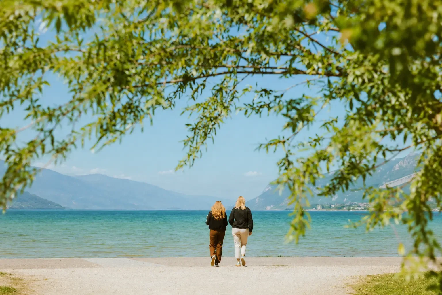 Deux personnes marchent sur une plage au bord d'un lac, avec des montagnes en arrière-plan et des branches d'arbre en premier plan